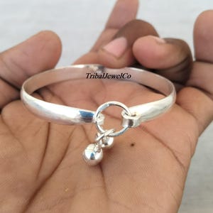 May include: A silver bangle with two small, dangling bells. The bracelet has a simple, rounded design. The text "TribalJewelCo" is visible on the bracelet.