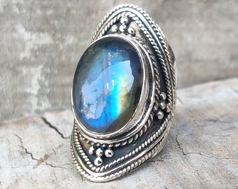 May include: A silver ring with a large oval-shaped blue and green labradorite gemstone set in a detailed silver bezel. The ring has a decorative design with intricate details.