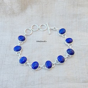 May include: A silver bracelet with a toggle clasp features oval-shaped lapis lazuli gemstones. The bracelet is on a light brown fabric background.