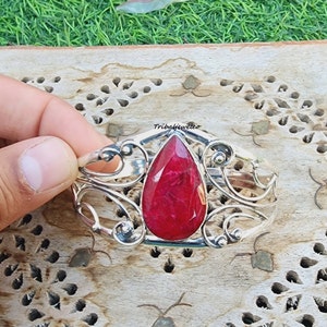 Può includere: Un braccialetto in argento con un designo ornato che presenta una grande pietra preziosa a forma di lacrima di colore rubino.