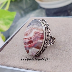 May include: A silver ring with an oval-shaped agate stone. The stone has a layered pattern of white, pink, and red. The ring has a decorative filigree design around the stone.