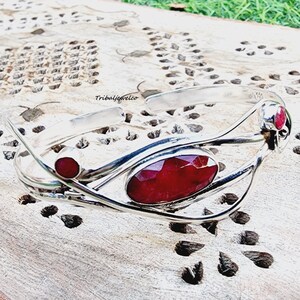 Peut inclure: Un bracelet en argent avec une grande pierre précieuse rouge et deux plus petites pierres rouges. Le bracelet est posé sur une surface en bois.