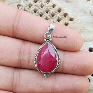 Könnte beinhalten: Ein tropfenförmiger Anhänger mit einem roten Edelstein, der in einen silbernen Rahmen gefasst ist. Der Anhänger hat einen zarten seilartigen Rand und eine kleine Öse oben. Der Text "TribalJewelCo" ist im Hintergrund sichtbar.