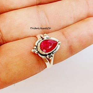Pode incluir: Um anel de prata com uma gema vermelha em forma de lágrima engastada em uma moldura de prata decorativa. O anel está em um dedo.