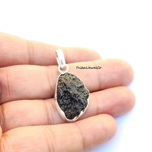 May include: A silver pendant featuring a dark brown, rough, and textured meteorite stone. The pendant is in the shape of an oval and has a silver bail.