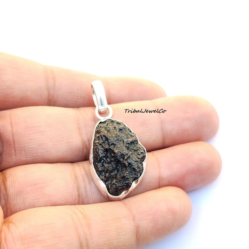 May include: A silver pendant featuring a dark brown, rough, and textured meteorite stone. The pendant is in the shape of an oval and has a silver bail.