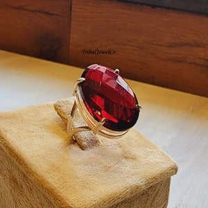 May include: A gold ring with a large, oval-shaped, red gemstone. The ring is on a beige velvet display box. The text "TribalJewelCo" is visible in the background.