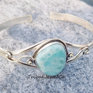 May include: A silver bangle bracelet with a light blue gemstone set in a silver bezel. The bracelet has a delicate, ornate design with two curved silver bands.