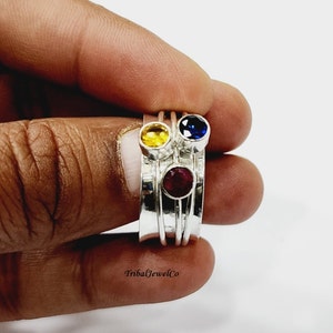 Peut inclure: Une bague en argent avec quatre pierres précieuses colorées. La bague a un design empilé avec une pierre jaune, rouge, bleue et une quatrième pierre qui n'est pas visible sur l'image. La bague est sur le doigt d'une personne.