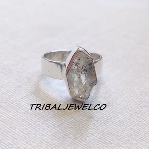 May include: A silver ring with a clear quartz crystal set in the center. The crystal is faceted and has a rough, natural look. The ring is a simple band with a polished finish.
