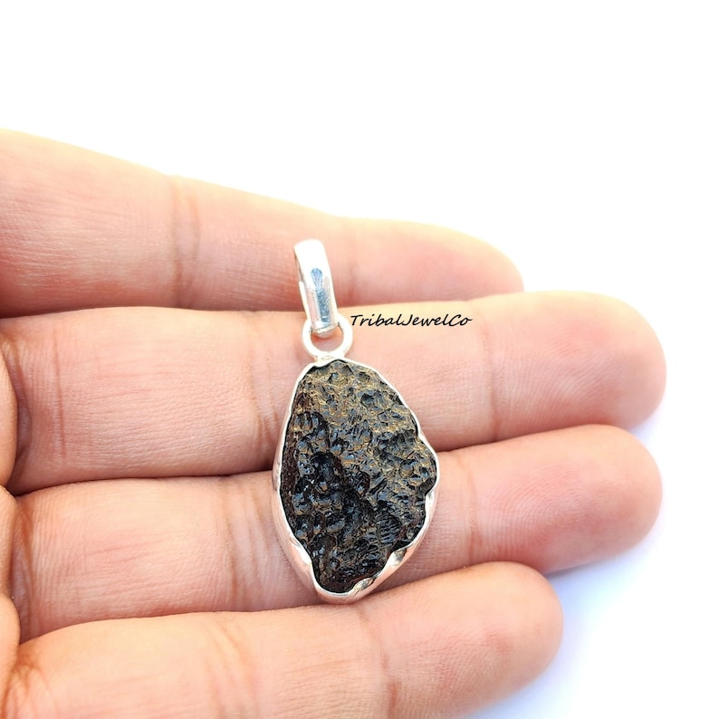 May include: A silver pendant featuring a dark brown, rough, and textured meteorite stone. The pendant is held in a hand.