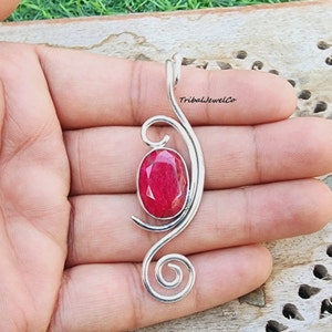 Könnte beinhalten: Ein silberner Anhänger mit einem roten Edelstein, der in einem Wirbeldesign eingefasst ist. Der Anhänger wird in einer Hand gehalten und der Text "TribalJewelCo" ist im Hintergrund sichtbar.