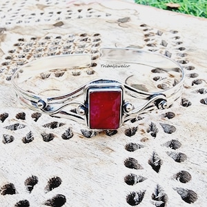 May include: A silver bangle bracelet featuring a red gemstone set in a square frame. The bracelet has a delicate, flowing design with two curved lines.