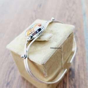 May include: Silver bangle bracelet with a colorful, multi-faceted gemstone set in the center. The bracelet is open and has a simple, elegant design.