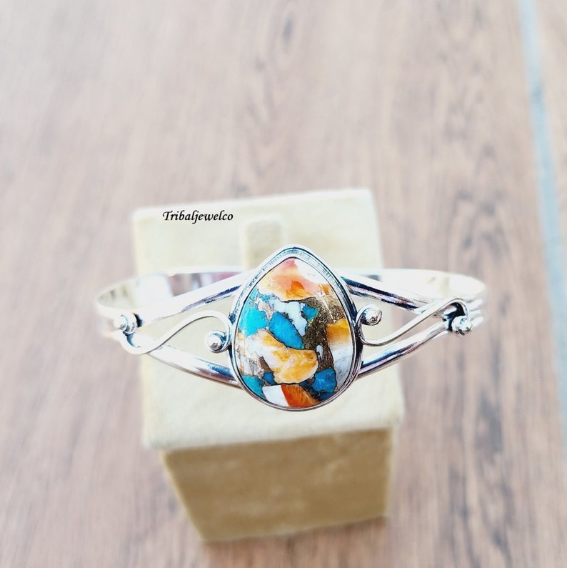 May include: A silver bangle bracelet with a teardrop-shaped turquoise stone set in the center. The stone has a mix of orange, blue, white, and brown colors.