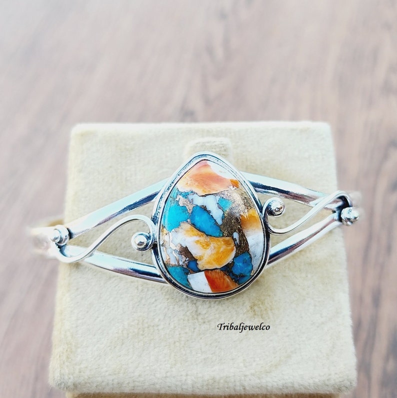 May include: A silver cuff bracelet with a teardrop-shaped turquoise stone set in the center. The stone is a mix of blue, green, orange, and white.