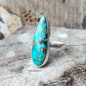 May include: A turquoise and gold teardrop-shaped ring set in sterling silver. The ring is on a wooden surface.