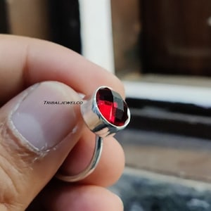 Puede incluir: Un anillo de plata con una piedra preciosa roja. El anillo está engastado en una banda simple y la piedra preciosa es redonda y facetada.