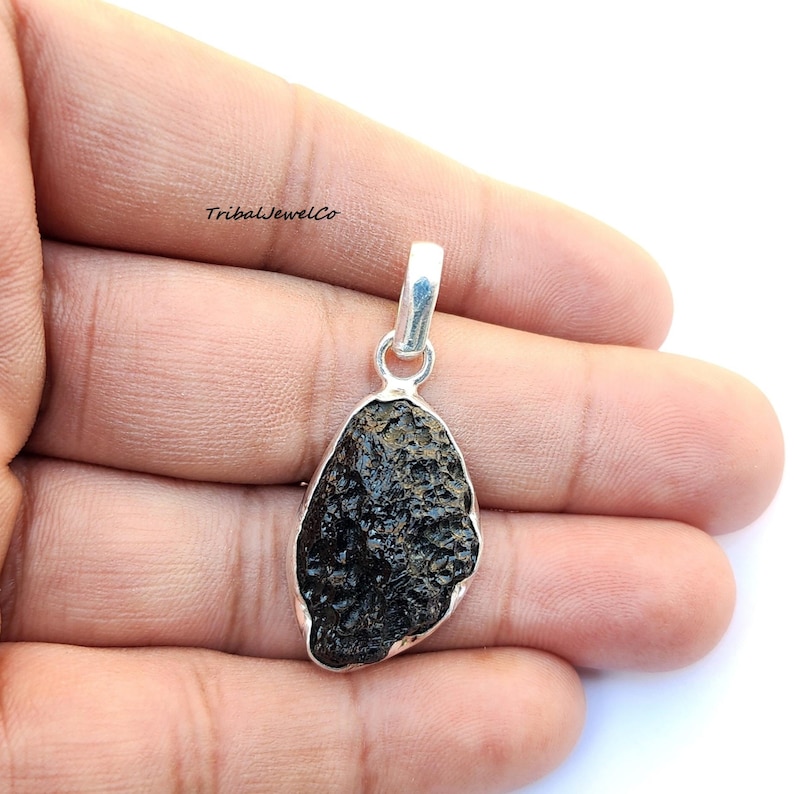 May include: A silver pendant featuring a dark gray, irregularly shaped meteorite with a smooth, polished surface. The pendant is attached to a silver bail.