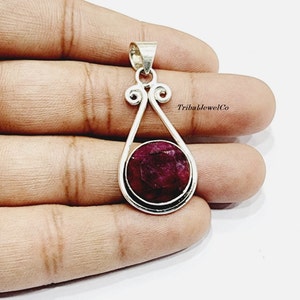 Può includere: Un ciondolo d'argento con una pietra preziosa di rubino rosso incastonata in un design delicato e ornato. Il ciondolo ha una forma a goccia con un rubino circolare al centro. TribalJewelCo è stampato sull'immagine.