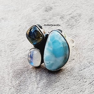 Könnte beinhalten: Ein silberner Ring mit einem großen blauen Edelstein, einem kleineren weißen Edelstein und einem kleineren dunkelgrauen Edelstein. Der Ring ist in einem Cluster-Design gefasst.