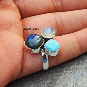 Op de afbeelding: Een zilveren ring met drie vierkante edelstenen. De edelstenen zijn lichtblauw, donkerblauw en wit. De ring wordt aan een vinger gedragen.
