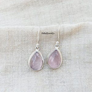 Puede incluir: Un par de pendientes de plata con piedras rosas en forma de lágrima. Los pendientes están sobre un fondo de tela blanca.