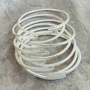 Puede incluir: Un conjunto de brazaletes plateados. Cada brazalete tiene un diseño en espiral con un extremo aplanado y texturizado. Las joyas se exhiben sobre una superficie gris texturizada. Los brazaletes tienen un diseño sencillo y elegante.