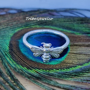 Può includere: Un anello d'argento con un designo di ape dettagliato. L'anello è su uno sfondo di piuma di pavone.