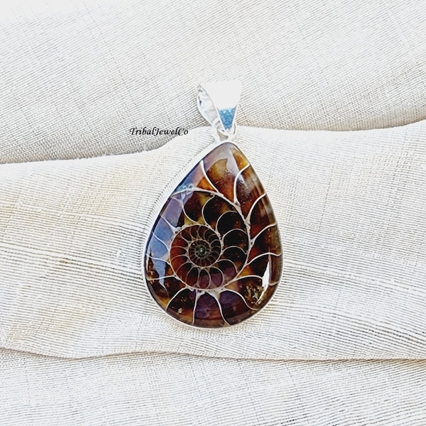 May include: A silver pendant featuring a teardrop-shaped ammonite fossil with a brown and black spiral pattern. The pendant is on a white fabric background.
