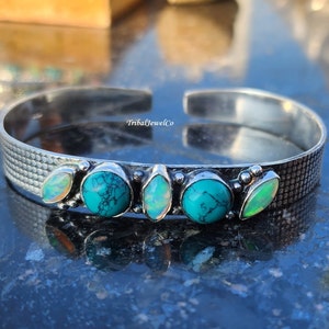 May include: Silver cuff bracelet with a textured surface and five stones. The stones are a mix of turquoise and opal, with a light blue and white colour scheme.