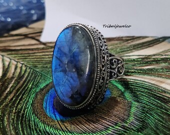 May include: A silver ring with a large oval blue labradorite gemstone set in a decorative silver band. The ring is on a peacock feather background.