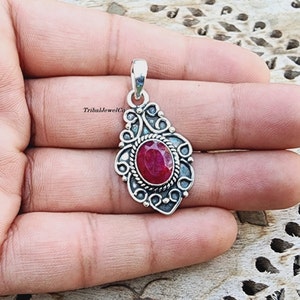 Op de afbeelding: Een zilveren hanger met een sierlijk ontwerp heeft een rode robijn edelsteen in het midden. De hanger wordt in een hand gehouden.