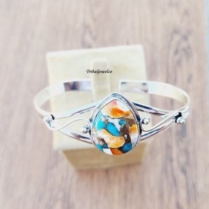 May include: A silver bangle bracelet with a teardrop-shaped turquoise and orange stone set in the center. The bracelet has a delicate, twisted design.