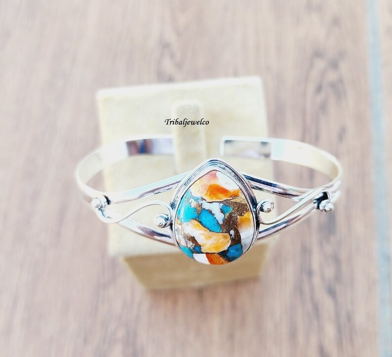 May include: A silver bangle bracelet with a teardrop-shaped turquoise and orange stone set in the center. The bracelet has a delicate, twisted design.