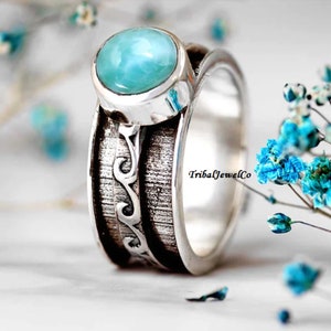 May include: A silver ring with a blue gemstone and a textured band with a wave design. The band is engraved with the text "TribalJewelCo".