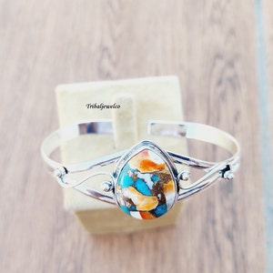 May include: A silver bangle bracelet with a teardrop-shaped turquoise and orange stone set in the center. The stone has a unique pattern of turquoise, orange, and white.