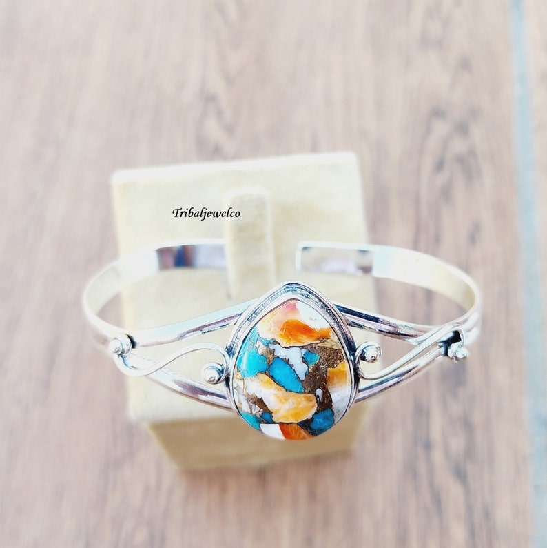 May include: A silver bangle bracelet with a teardrop-shaped turquoise and orange stone set in the center. The stone has a unique pattern of turquoise, orange, and white.