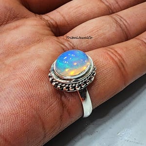 May include: A silver ring with an oval-shaped opal gemstone. The opal is iridescent with blue, green, and yellow hues. The ring has a beaded and twisted band.