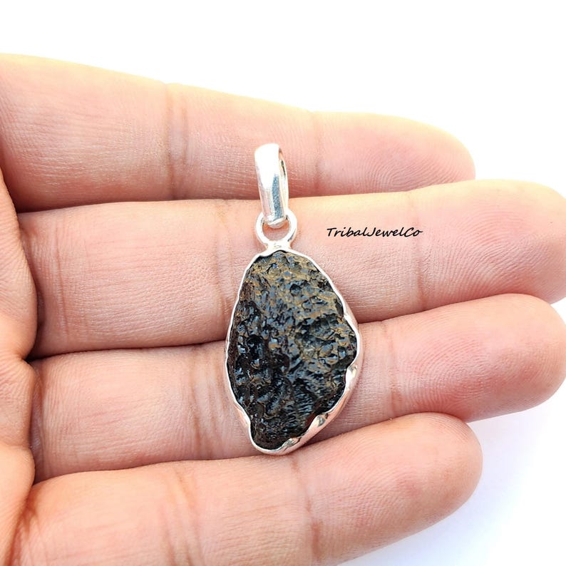 May include: A silver pendant featuring a dark grey, rough, and textured meteorite stone. The pendant is oval-shaped and has a silver bail.