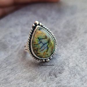 May include: A silver ring with a pear-shaped, multi-colored gemstone. The stone features shades of green, blue, and orange with a unique pattern. The ring has a twisted rope design around the stone and small beaded accents. The brand name "TribalJewelCo" is visible.