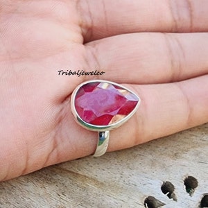 May include: A silver ring with a large, faceted, teardrop-shaped red gemstone. The ring is on a person's finger.
