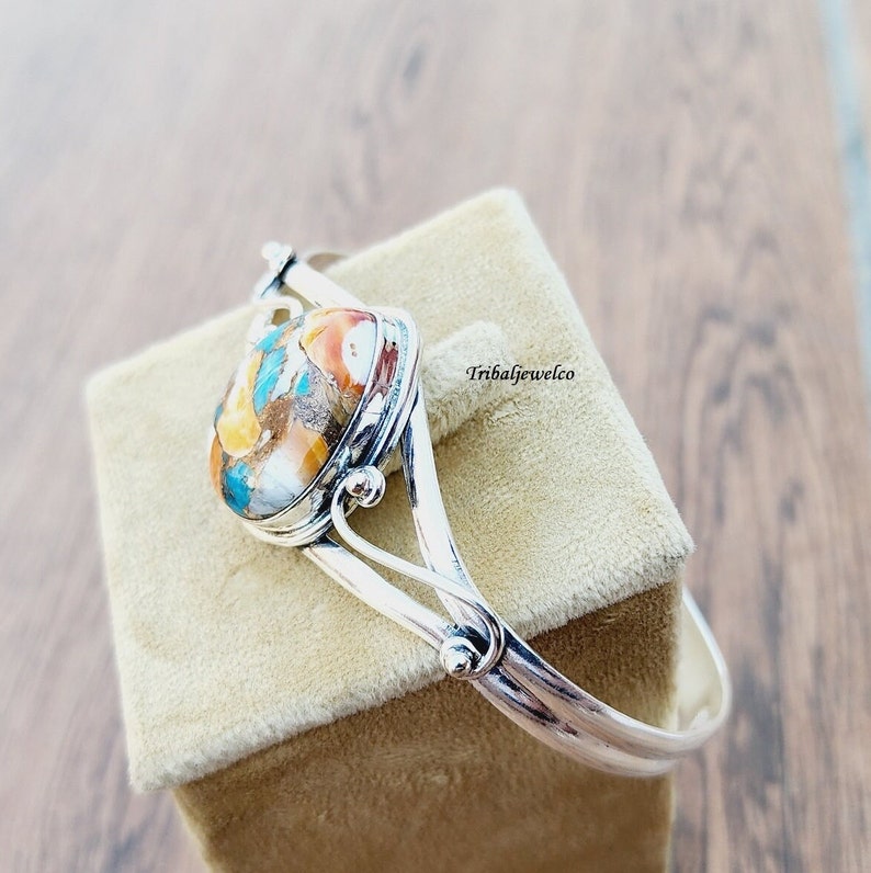 May include: A silver bangle bracelet with a large oval-shaped gemstone set in the center. The gemstone is a mix of turquoise, brown, orange, and white colors. The bracelet is on a beige velvet box.