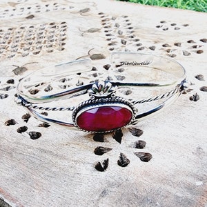 Könnte beinhalten: Silbernes Armreif-Armband mit einem roten Edelstein und einem gedrehten Drahtdesign. Das Armband hat ein kronenförmiges Detail oben.