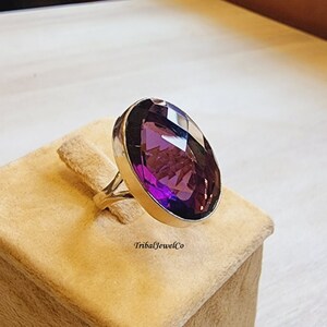 May include: A silver ring featuring a large, oval-shaped amethyst gemstone. The ring is displayed on a white velvet stand.
