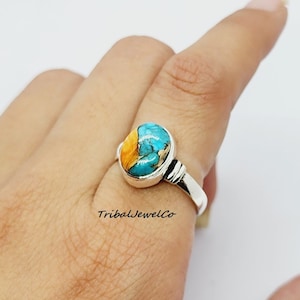 Puede incluir: Un anillo de plata con una piedra ovalada azul y naranja. El anillo está en un dedo.