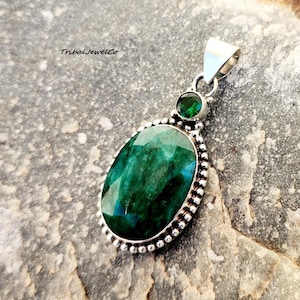 Pode incluir: Um pingente de prata com uma grande pedra preciosa oval verde cercada por uma borda de contas. Uma pedra preciosa verde menor está engastada acima da maior.
