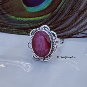 May include: A silver ring featuring a pink oval gemstone set within a decorative floral design. The ring is displayed on a white surface with a peacock feather in the background.