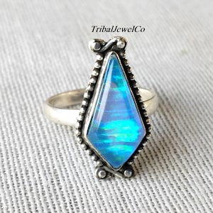 Op de afbeelding: Een zilveren ring met een grote, iriserende blauwe edelsteen in een langwerpige diamantvorm. De edelsteen is gezet in een zilveren bezel met een kralenrand. De ring heeft een eenvoudige band en wordt gepresenteerd op een getextureerd, lichtgrijs oppervlak.