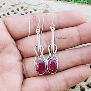 May include: A pair of silver dangle earrings with red oval gemstones. The earrings feature a delicate knot design and are accented with a silver hook.